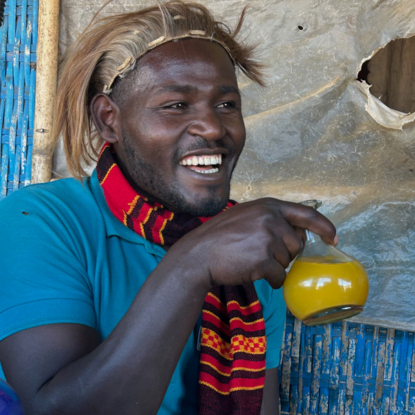 Kia, Ethiopian cultural guide with traditional hairstyle and colorful clothing, smiling while holding glass of golden honey wine (tej)